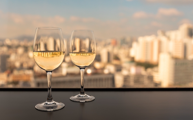 Two glasses of white wine on a table with a scenic view of the Seoul Korea city skyline under warm evening light, symbolizing romance, relaxation, and urban lifestyle.
