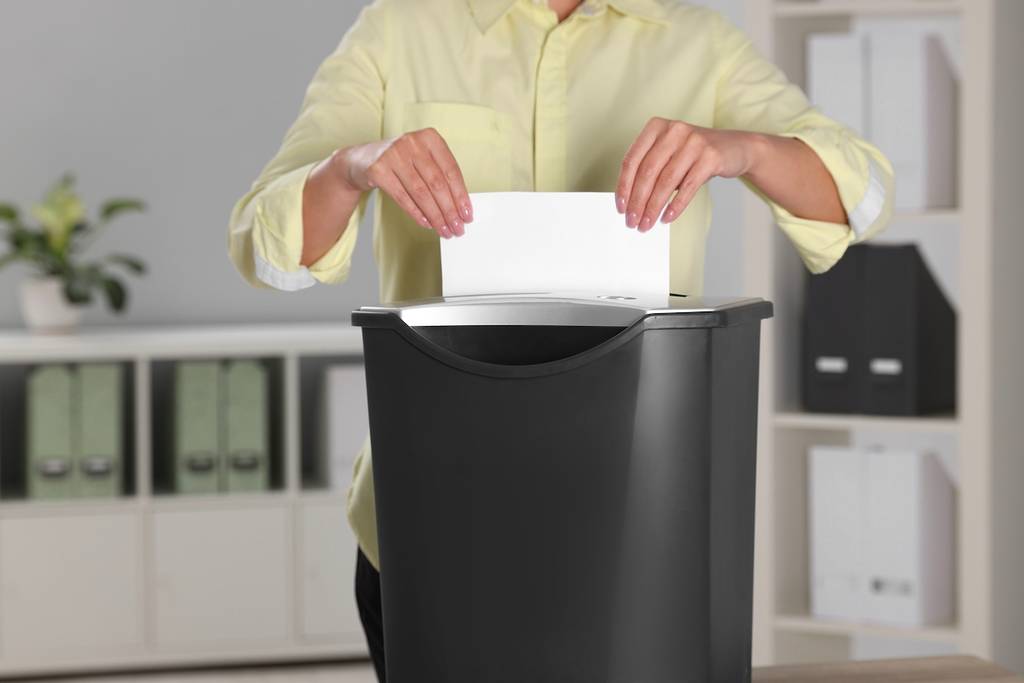 Woman destroying sheet of paper with shredder in office, closeup