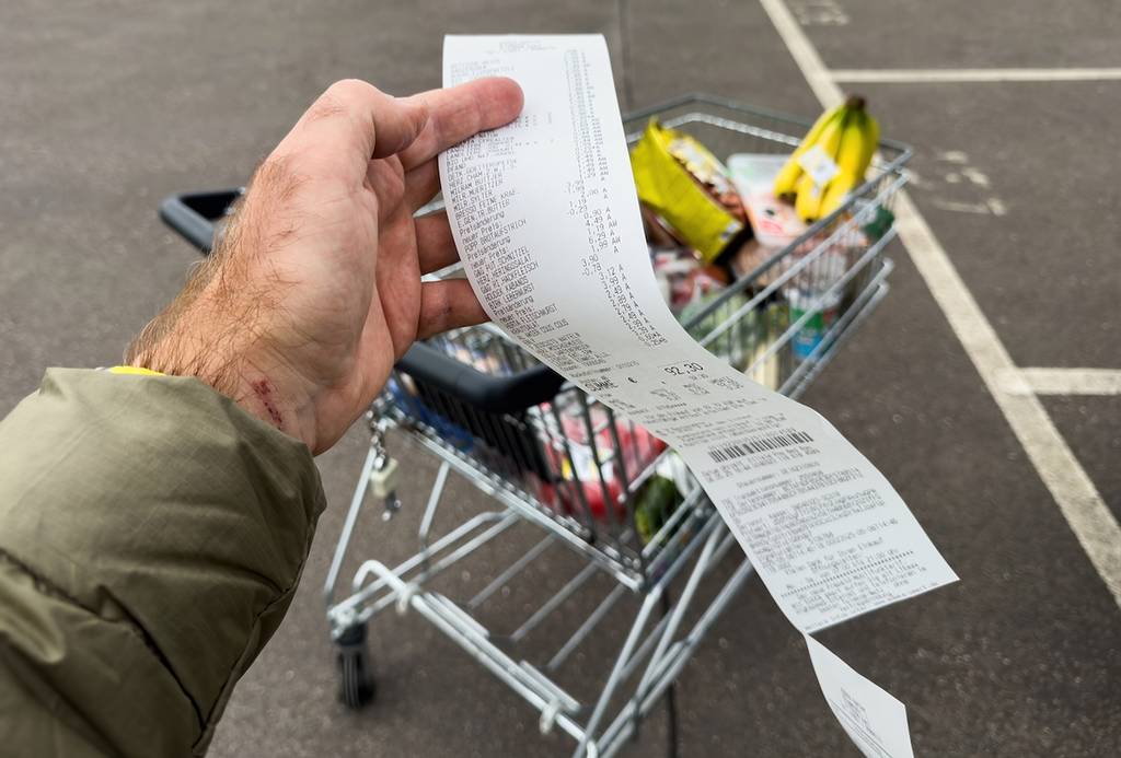 Frankfurt, Germany - May 6, 2025: A person holds a long grocery receipt in front of a full shopping cart in a nearly empty parking lot showing a large number of purchased items