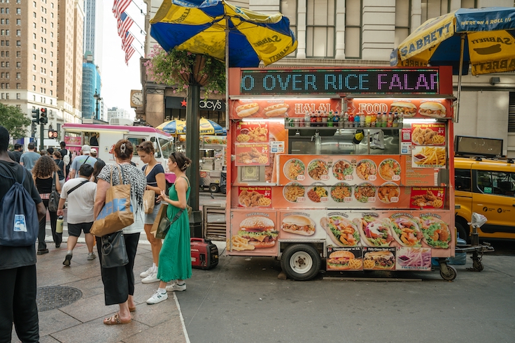 Nueva York, Nueva York, EE. UU. - 29/07/2024: Un colorido puesto de comida halal atiende a los clientes en una concurrida calle de Nueva York. La gente hace cola rodeada de tráfico, taxis amarillos y los emblemáticos Sabrett.