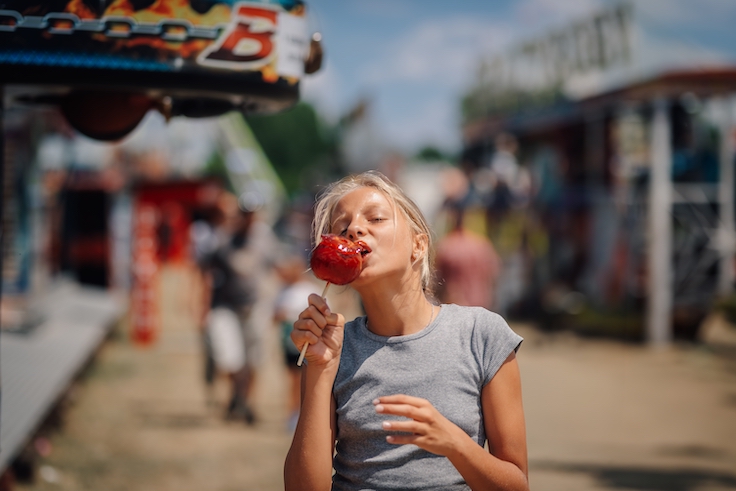 Blonde girl savoring a delicious caramel apple at a vibrant funfair, embracing the joy of a sunny summer day outdoors