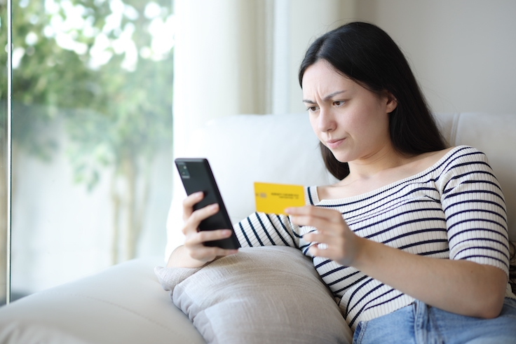 Una mujer asiática molesta comprando por internet con el móvil y la tarjeta de crédito, sentada en el sofá de su casa