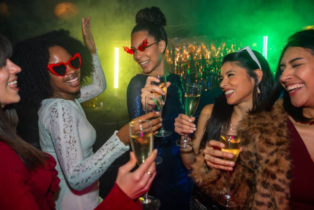 Diverse group of smiling young women toasting with champagne in a vibrant nightclub, celebrating a special occasion with colorful lights, dancing, and joyful togetherness