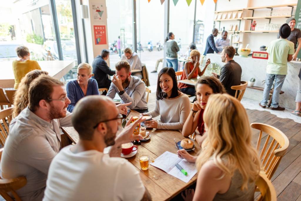 Un grupo variado de gente que disfruta del café y la charla en un animado ambiente de cafetería. Se sientan alrededor de mesas de madera en un ambiente luminoso y acogedor.