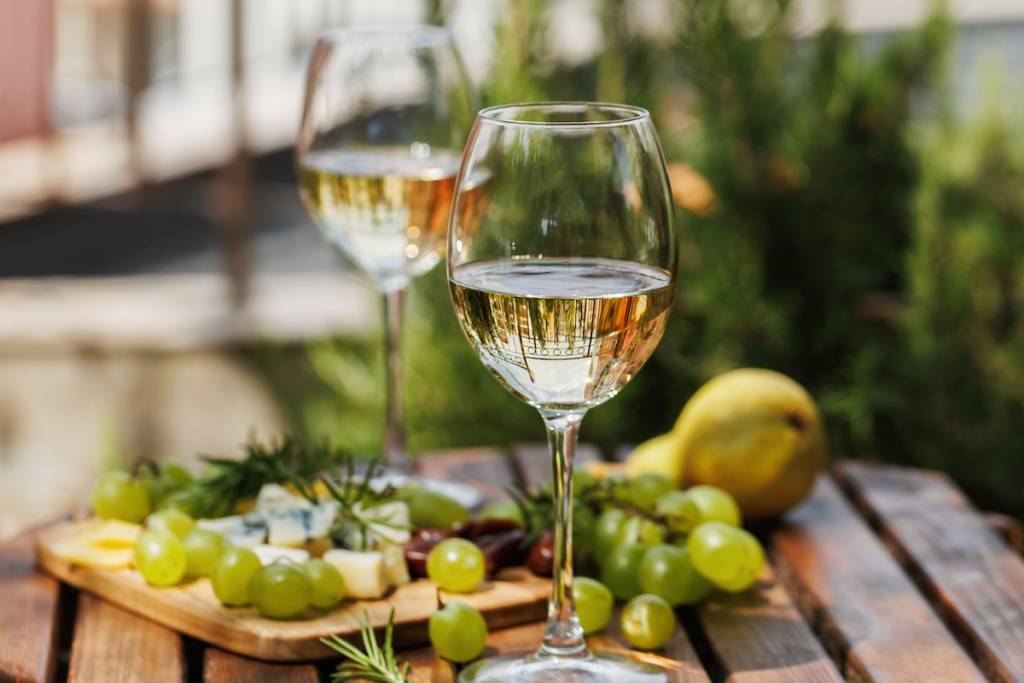 Una elegante mesa en el jardín con dos copas de vino blanco, uvas verdes, una pera y una tabla de quesos bajo la luz del sol