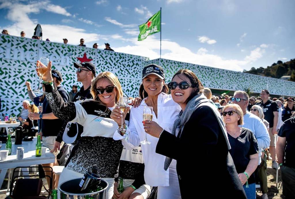 Three women attending SailGP pose for a photo with drinks in their hands.