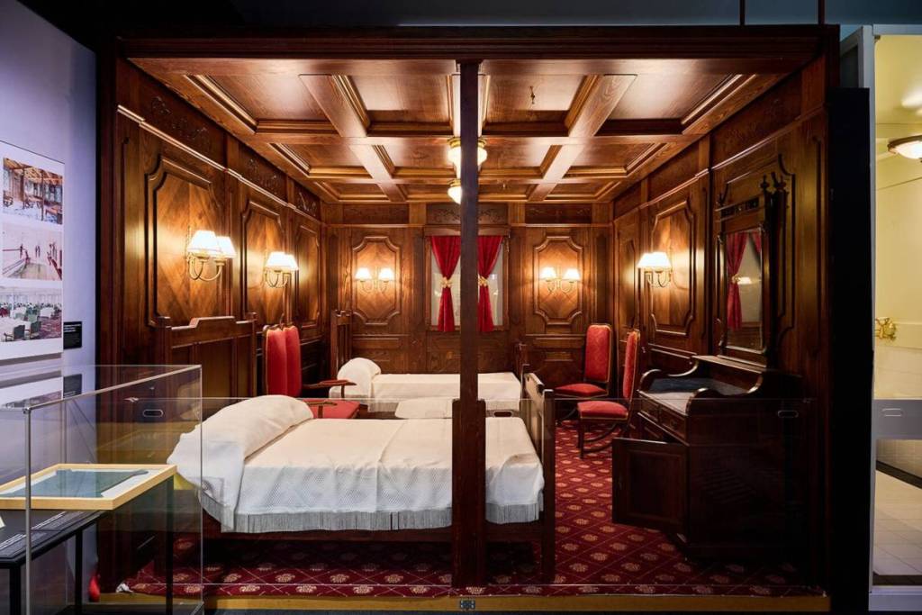 A recreation of the upper class cabins aboard the Titanic including beds, lit wall lamps, a writing desk and upholstered chairs.