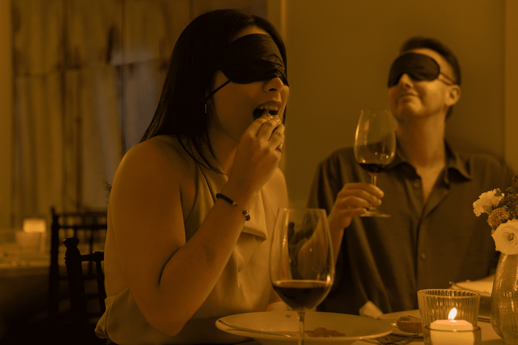 A woman who is blindfolded eating food from her plate while her partner drinks wine