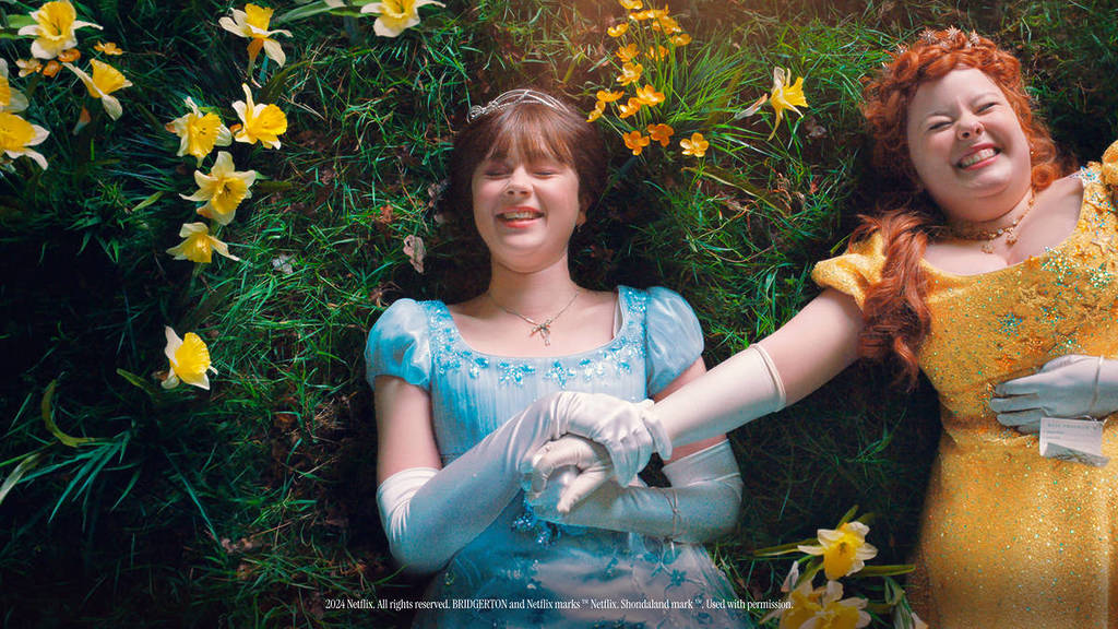 Deux protagonistes de la série Bridgerton sont allongées en riant dans une verte prairie avec des fleurs autour d'elles.