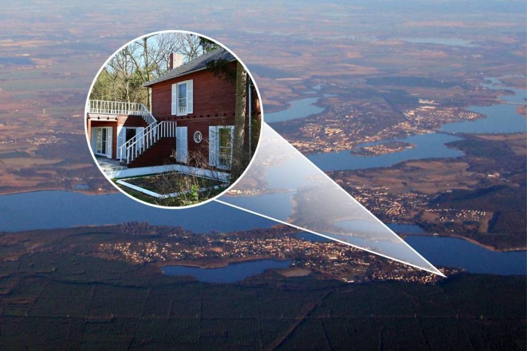 Vista aerea dell’ampio paesaggio lacustre e boschivo intorno a Caputh. Una lente di ingrandimento grafica ingrandisce un punto sulla riva e mostra in dettaglio la casa di legno di Einstein.