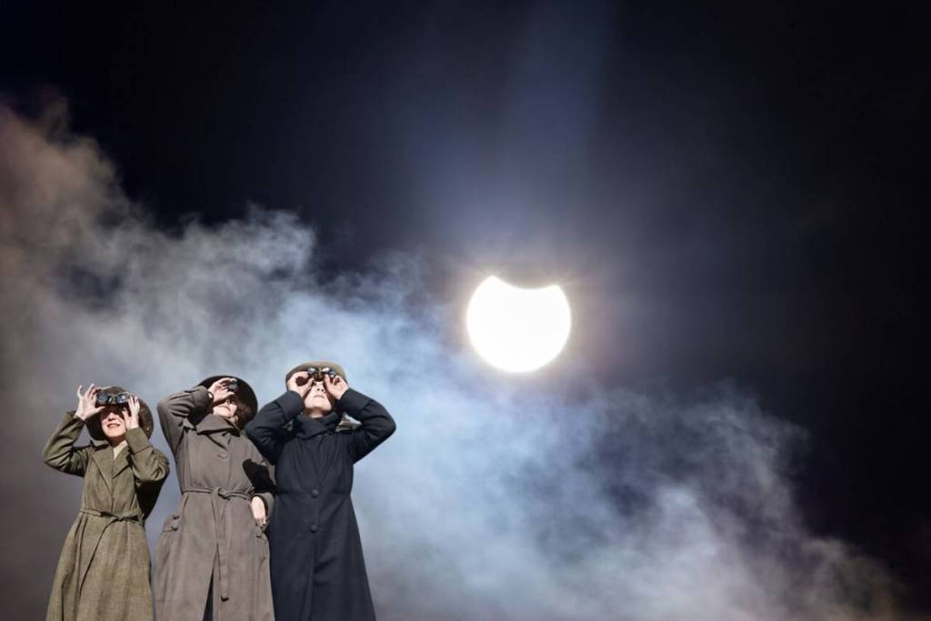 Tre persone in abiti vintage (cappotti invernali, cappelli) guardano in alto attraverso un binocolo per osservare un’eclissi solare parziale (una falce) che si staglia contro un cielo notturno scuro e nuvoloso.