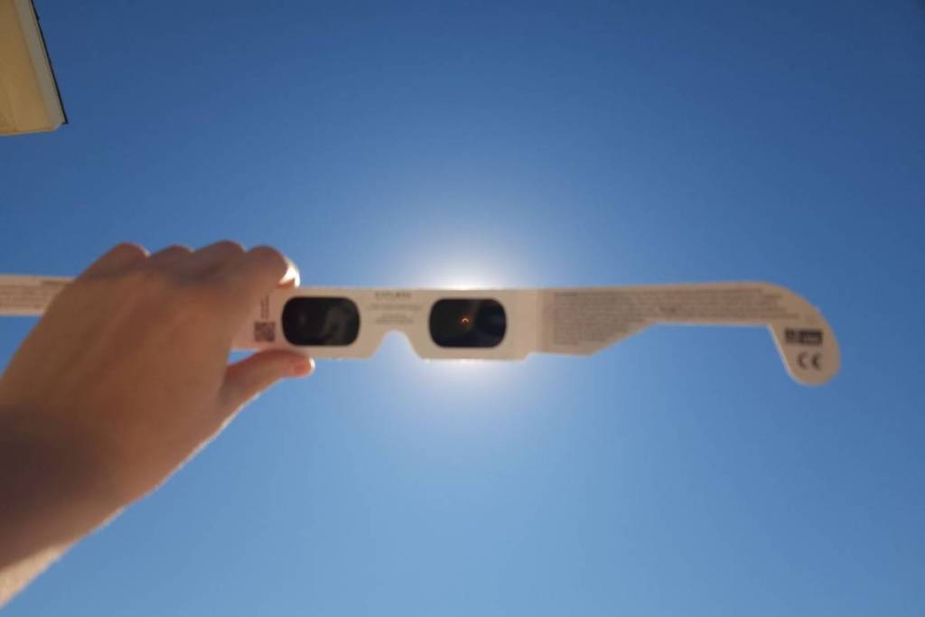 Una mano tiene un paio di occhiali per l’eclissi di carta bianca contro un cielo blu splendente. Attraverso le lenti scure degli occhiali si vede un sole parzialmente eclissato (una piccola falce).