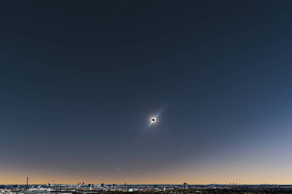 Una foto panoramica a grandangolo mostra lo skyline di Berlino in lontananza al tramonto. In alto, nel cielo blu intenso, si stacca un’eclissi solare con una corona luminosa, affiancata da una stella, su uno sfondo arancione che avvolge la città.