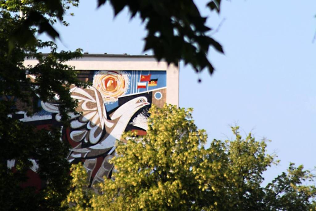 Ein farbenprächtiges Wandmosaik aus der DDR-Zeit an einer Hausfassade, das teilweise durch grüne Baumblätter verdeckt wird. Das Kunstwerk zeigt eine große weiße Friedenstaube, eine strahlende Sonne sowie die Flaggen der Sowjetunion und der DDR.
