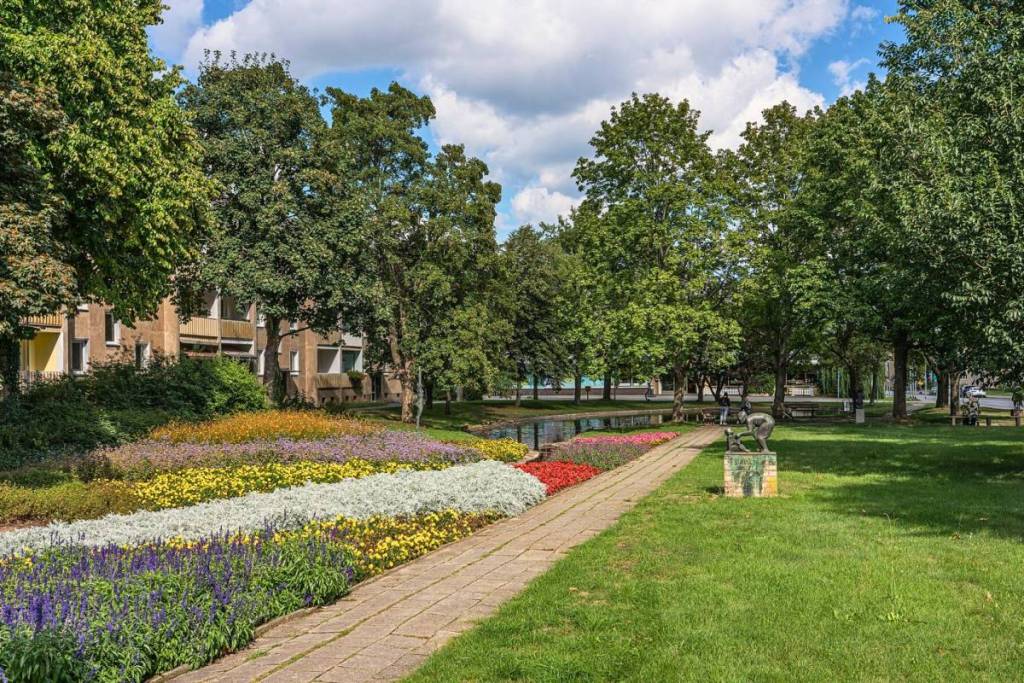 Eine gepflegte, grüne Parkanlage in einem Wohnkomplex in Eisenhüttenstadt. Im Vordergrund blühen farbenfrohe Blumenbeete neben einem gepflasterten Spazierweg, der an einer kleinen Bronzestatue und großen Bäumen vorbeiführt.