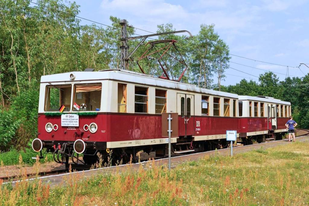 Die historische Buckower Kleinbahn im Nostalgie-Look: Ein rot-cremefarbener, elektrischer Triebwagen der Museumsbahn steht an einem sonnigen Tag umgeben von viel Grün an einem ländlichen Haltepunkt.