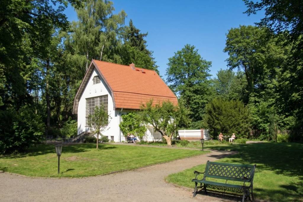 Das weiße Brecht-Weigel-Haus mit seinem roten Ziegeldach, malerisch eingebettet in einen weitläufigen, sonnigen Garten mit altem Baumbestand. Im Vordergrund steht eine einladende grüne Parkbank auf einer Wiese.
