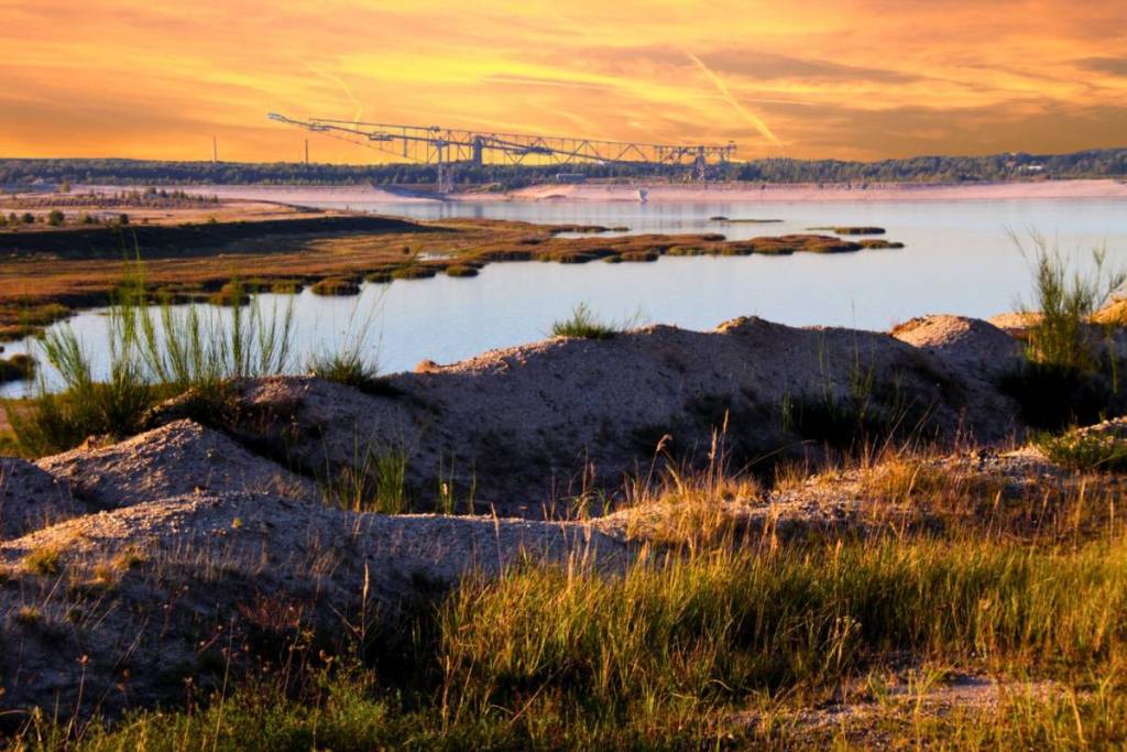 Scena paesaggistica nella Lusazia: in primo piano colline sabbiose ed erba secca, dietro lo specchio d’acqua del lago inondato e sullo sfondo la sagoma della miniera visitabile F60 di Lichterfeld.
