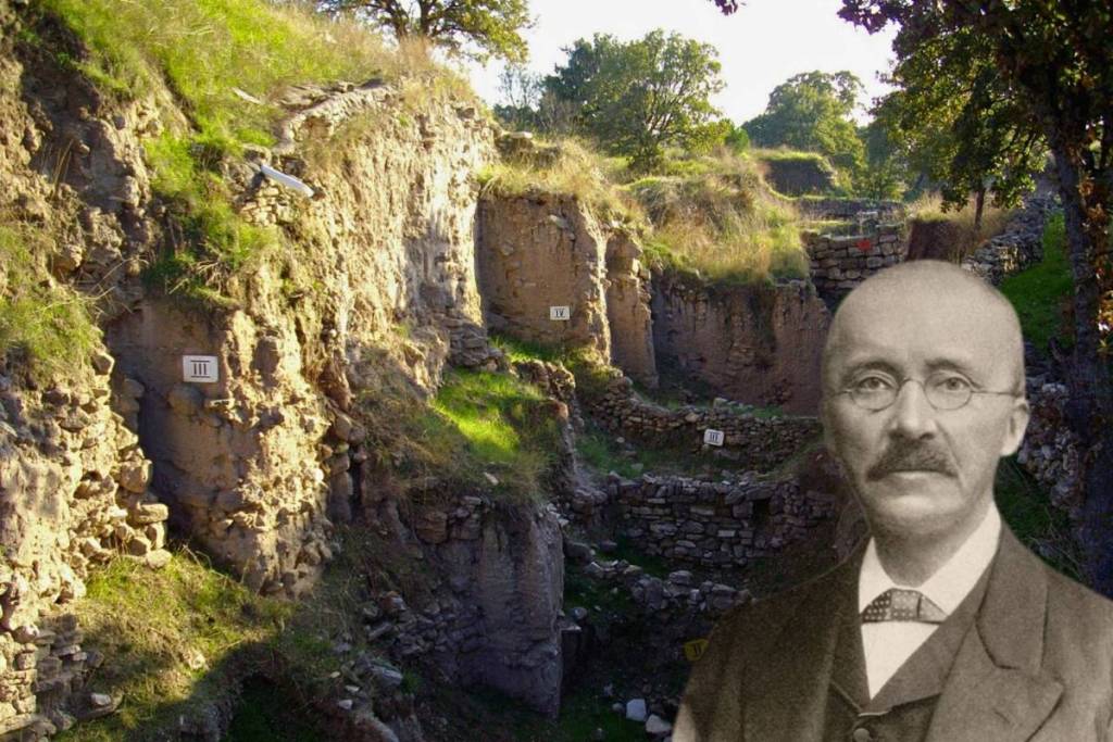 Primo piano del sito archeologico di Troia sulla collina di Hisarlik con profondi fossati e strati murari visibili, contrassegnati dagli strati Troia IV e Troia III. In basso a destra è inserito un ritratto storico di Heinrich Schliemann.