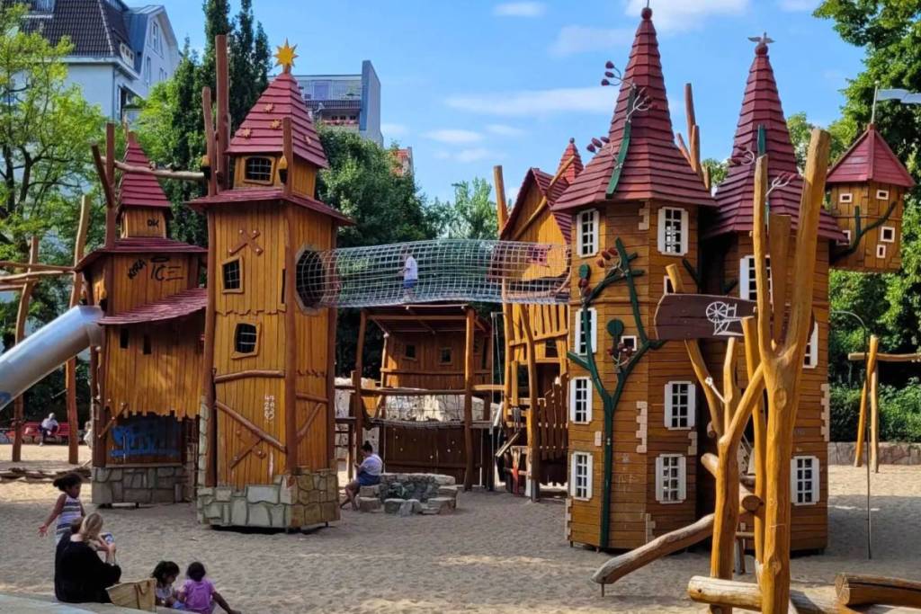 A wide-angle shot of the fairy-tale playground in Heinrich-Lassen-Park, Berlin Schöneberg, featuring the large, intricately detailed wooden castle, a metal slide, climbing bridges, and children playing in the sand.