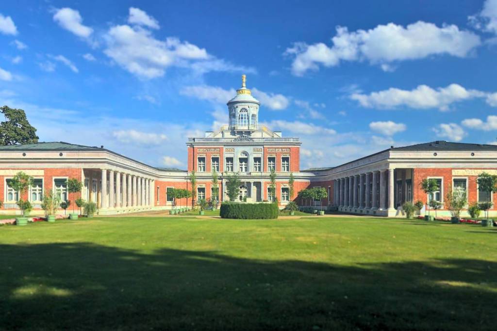 Vista general soleada del Marmorpalais, de estilo neoclásico temprano, en el Neuer Garten de Potsdam, con su amplia pradera y su llamativa fachada de mármol rojo de Silesia.