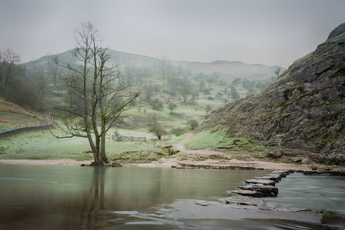 Stepping stones across river in misty winter valley landscape in Dovedale