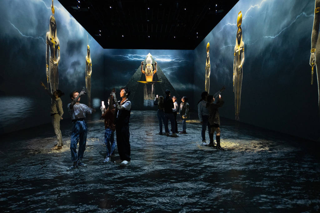 guests of the exhibition exploring the immersive room in a dark background, taking photos, walking around as the image constantly changes.