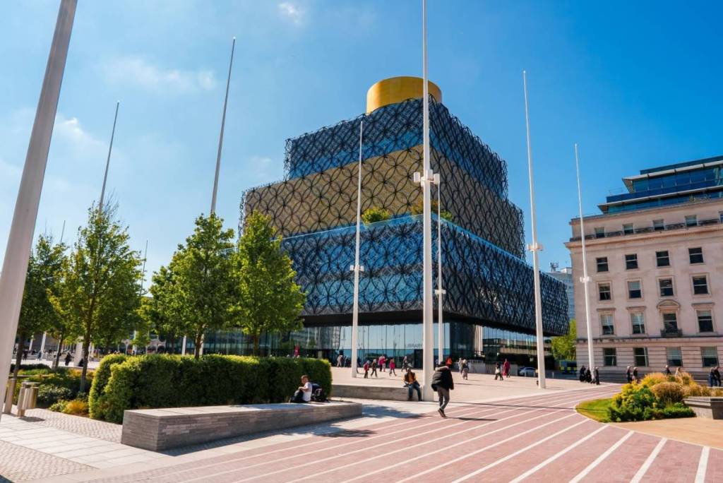 Birmingham, UK. May 5, 2023. A modern Birmingham plaza boasts a dark building with a chainmail pattern, golden peak, white poles, and folks basking in the sun, near a classic white facade.