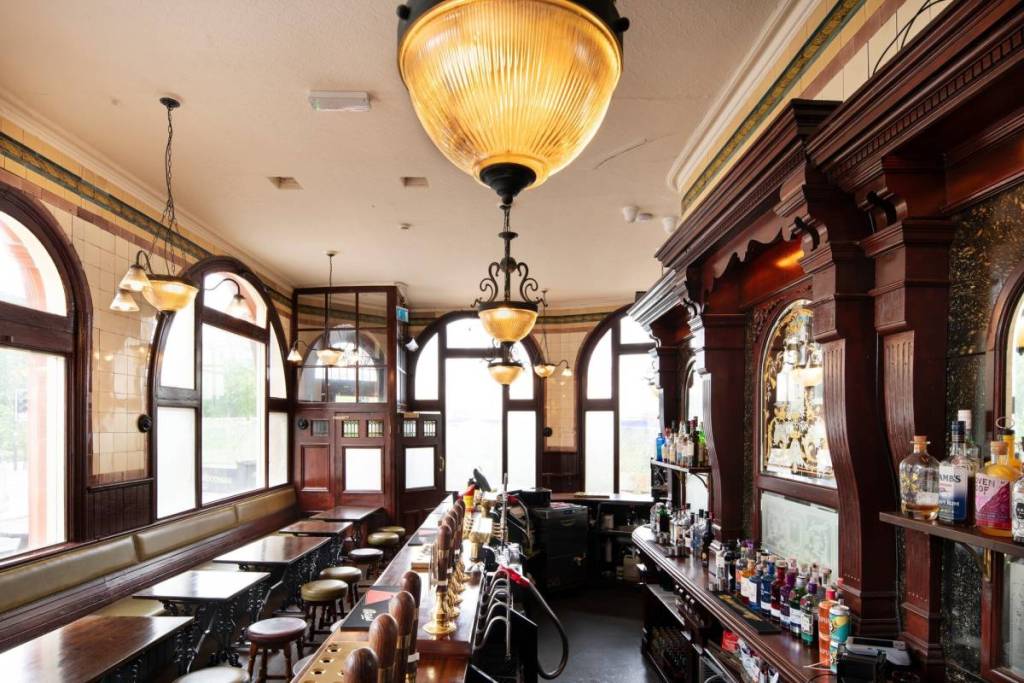 The interior of the Woodman, it features wooden bar, historic lighting, and small tables by arched windows.