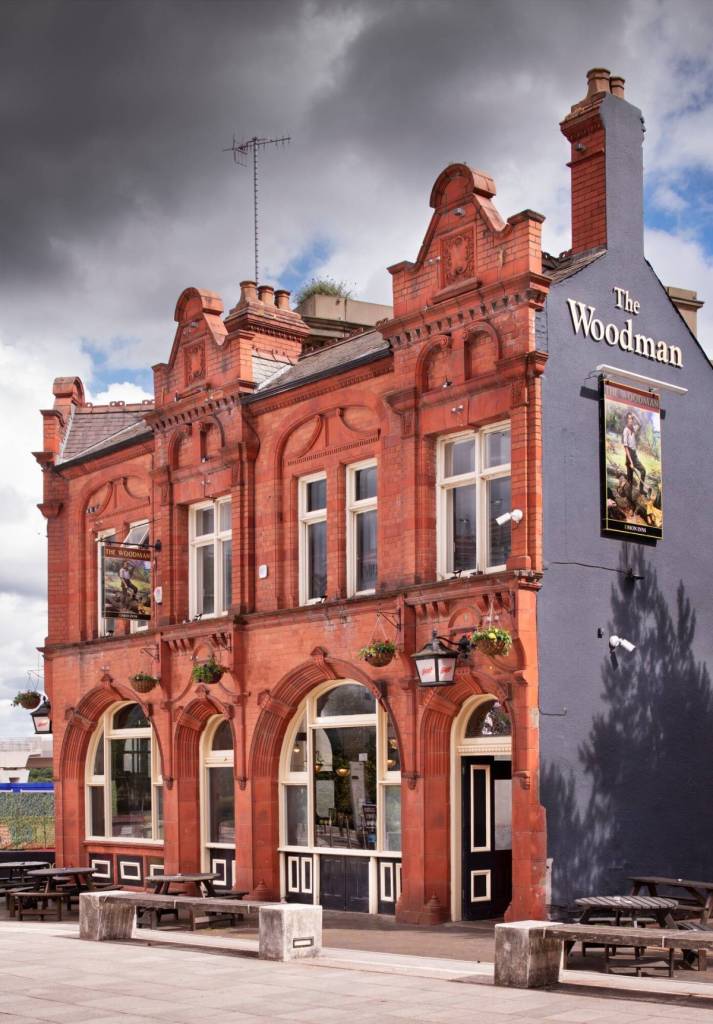 Photo of the Woodman in Birmingham by James O. Davies of Historic England. The Woodman won the Historic England Conservation category in CAMRA's Pub Design Awards 2026. The exterior of the Woodman, a traditional red-brick British pub with arched windows and outdoor picnic tables under a cloudy sky. The right hand side is painted black with signage.