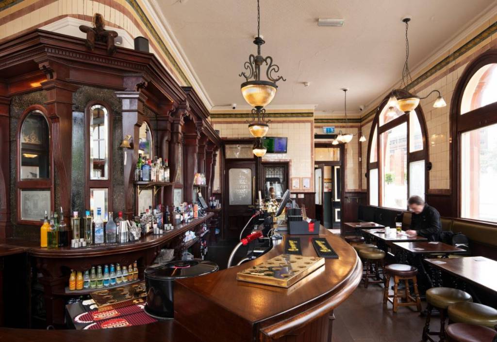 The interior of the Woodman, it features wooden bar, historic lighting, and small tables by arched windows.

