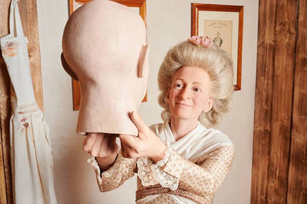 a waxwork of the founder of Madame Tussauds, Marie Tussaud, who is wearing a dress with lace detailing and holding up a waxwork head to study it