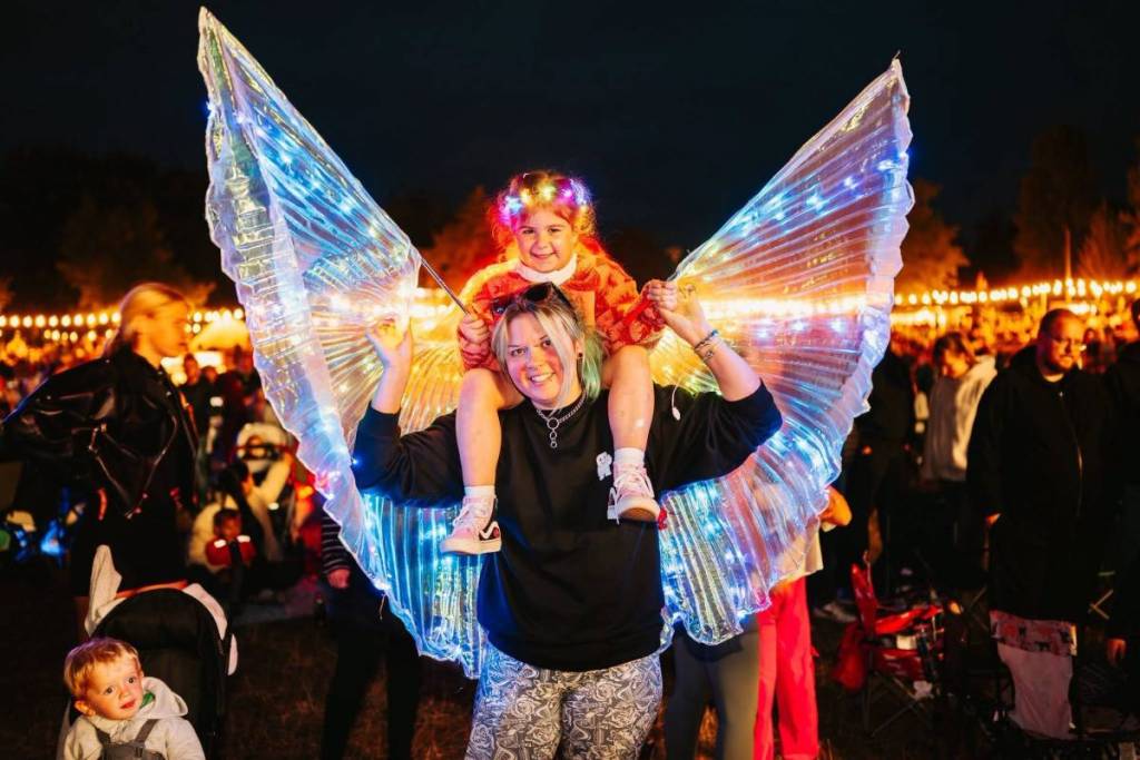 A woman wearing glowing angel wings with a child on her shoulders
