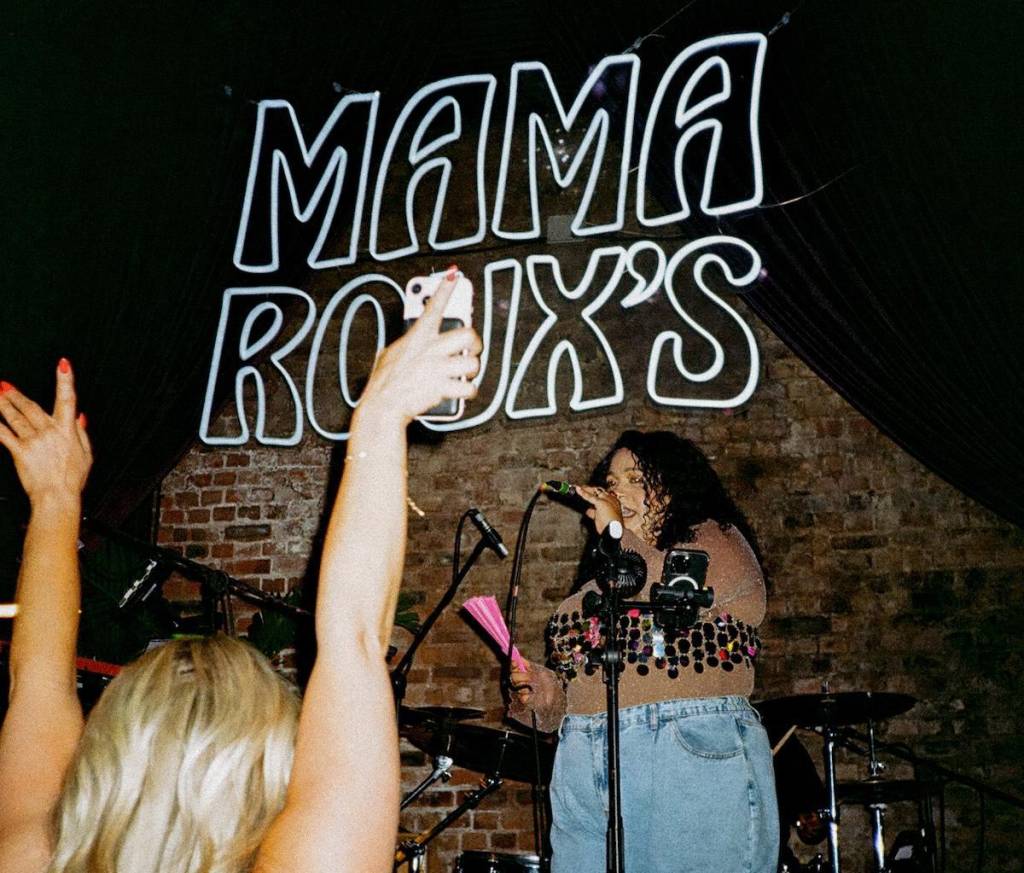 a woman stands on stage singing, a sign saying mama roux's is on the wall behind her