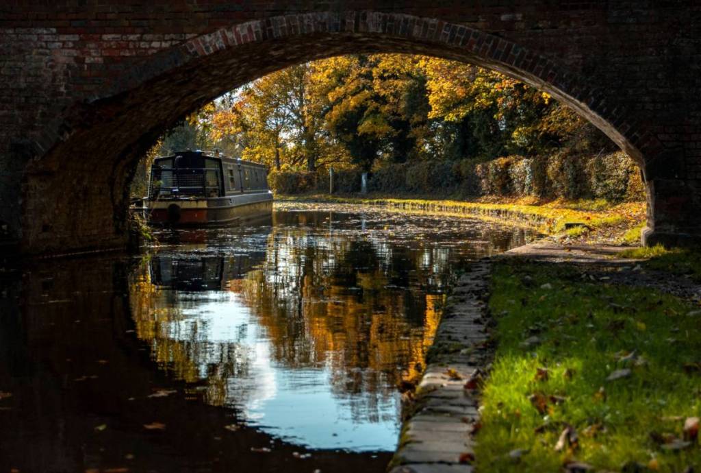 a view of a narrow boat under the arch of a brick foot bridge reflected into the still water of the canal, with autumn colours in the trees

