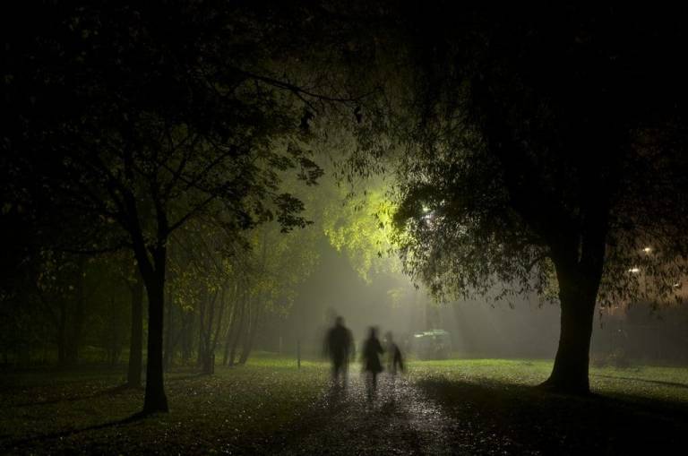 A wooded area at night, with s single light source illuminating the trees, while three shadowy people walk between the trees