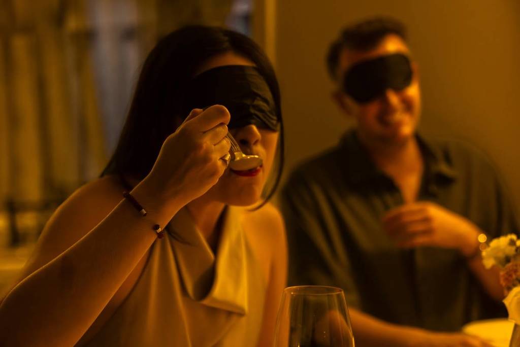 A blindfolded woman eating at a dimly lit restaurant during an evening of Dining in the Dark