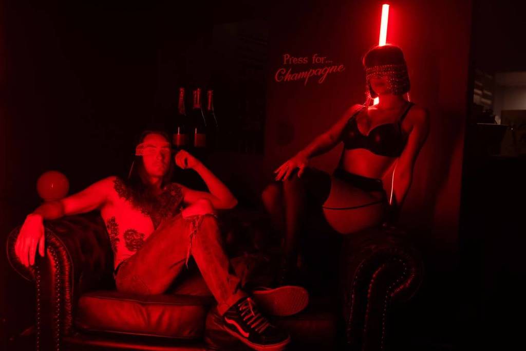 A man and a woman pose in a dark room with red lighting. The man sits on a couch and the woman stands. They are wearing blindfolds to promote Late Night Vice.