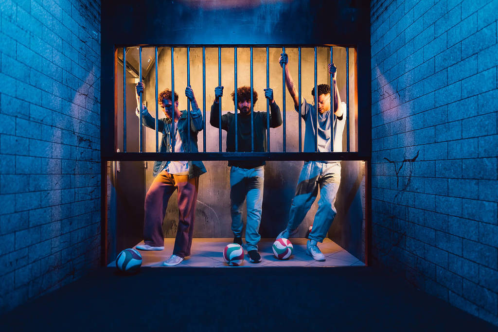 Three young men hold on to a barred gate and prepare to kick soccer balls into a cell at Prison Island.