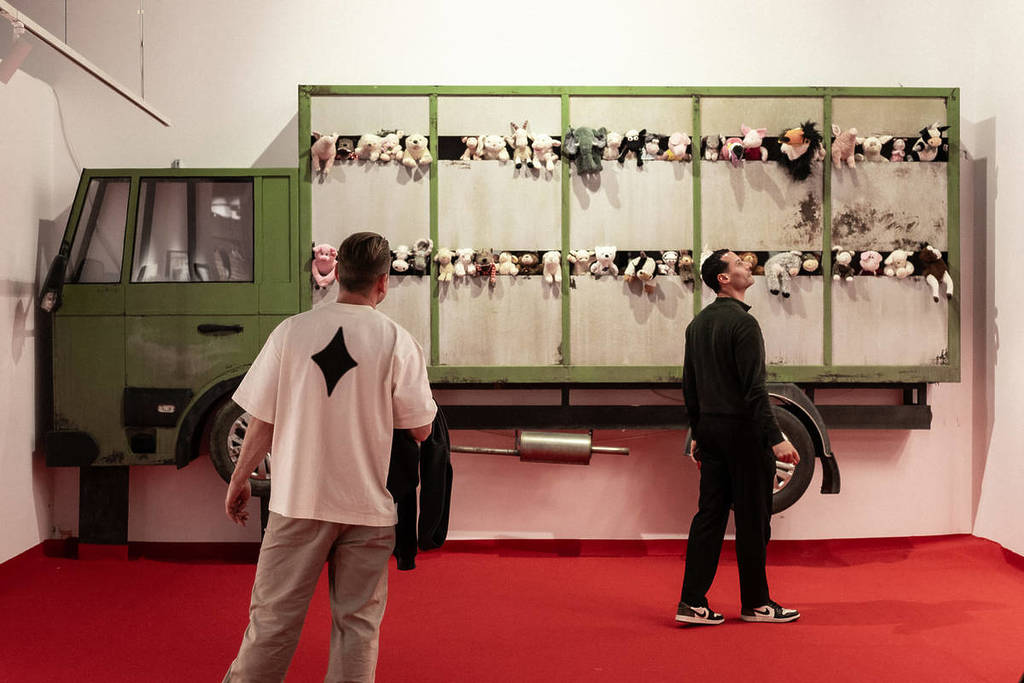 Two men observe an installation at The Art of Banksy: Chapter 2. The frame of a green truck is mounted on a wall, filled with stuffed animal toys.
