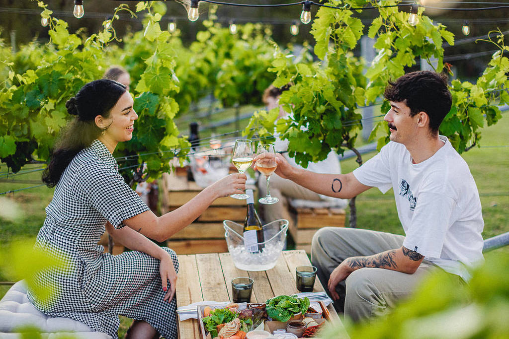 A young man and woman sitting at a low table in a vineyard cheers with their wine glasses.