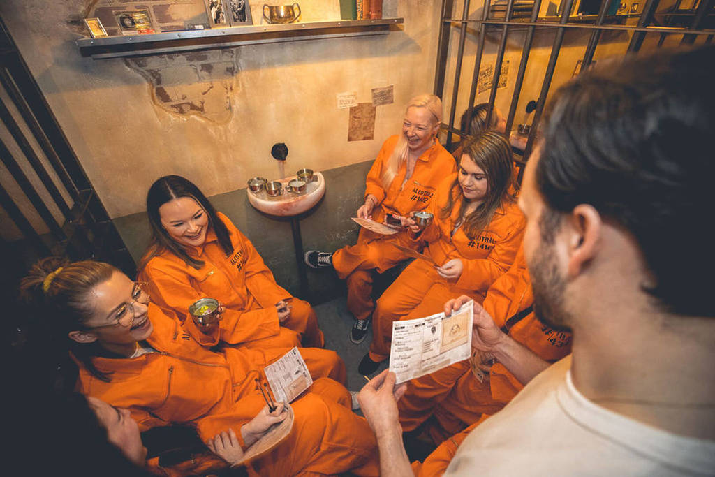 A group of friends in orange jumpsuits laugh together holding cocktails inside a prison cell at Alcotraz.