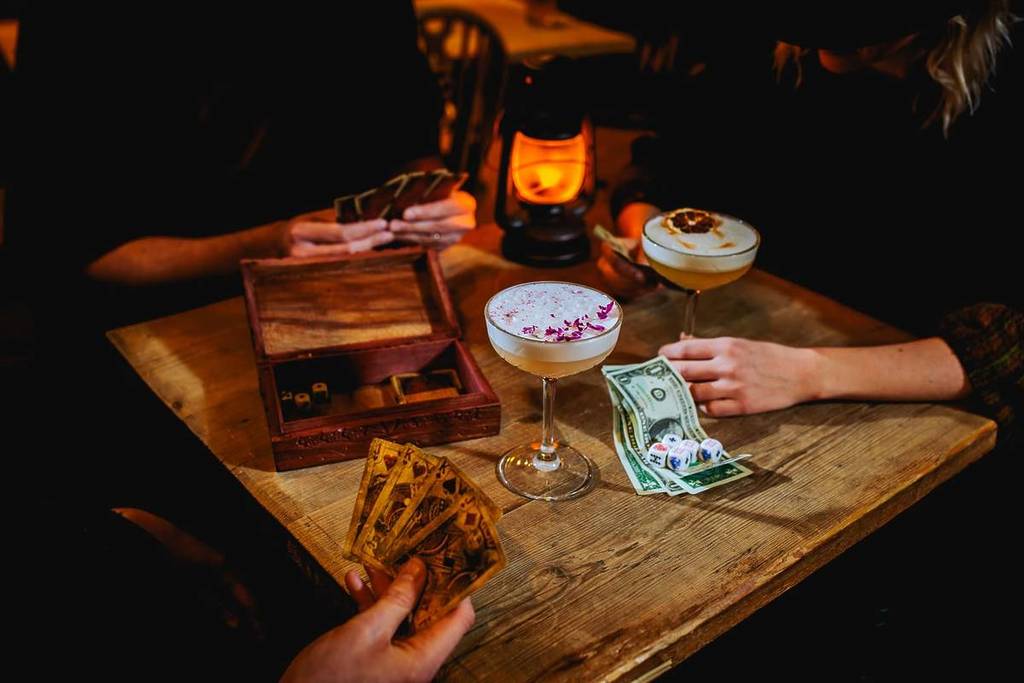 A close-up shot of a table with cocktails, a lamp and money on it as three people play cards at Moonshine Saloon.