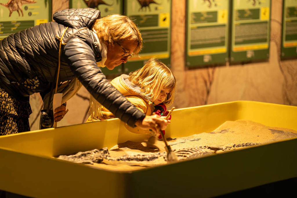 A mother and her young daughter lean over a sandbox and brush sand off a fossil at Dinos Alive.