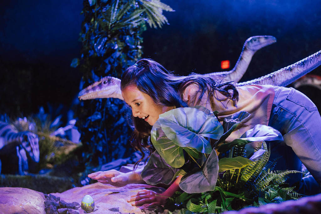 A young girl leans close to peer at a dinosaur egg at Dinos Alive. She is surrounded by small dinosaurs and green shrubbery.