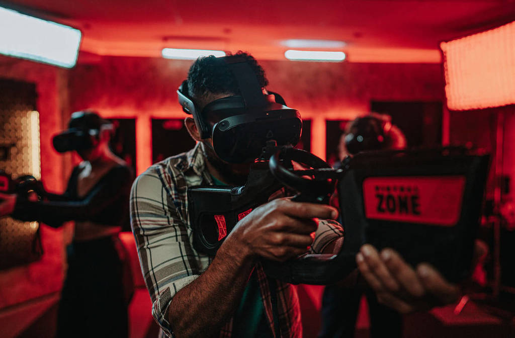 A close up shot of a man wearing a VR headset at Virtual Zone, pointing his VR gun. He is inside a room with red lighting, his two friends behind him.