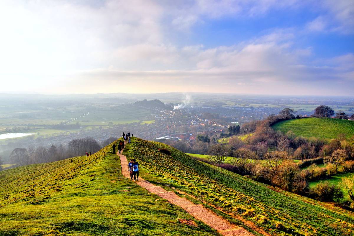 Glastonbury, UK - December 30, 2019: Tourists walking on the path to Glastonbury Tor, in a golden winter sunset with clear skies, view on the city and fields on the background