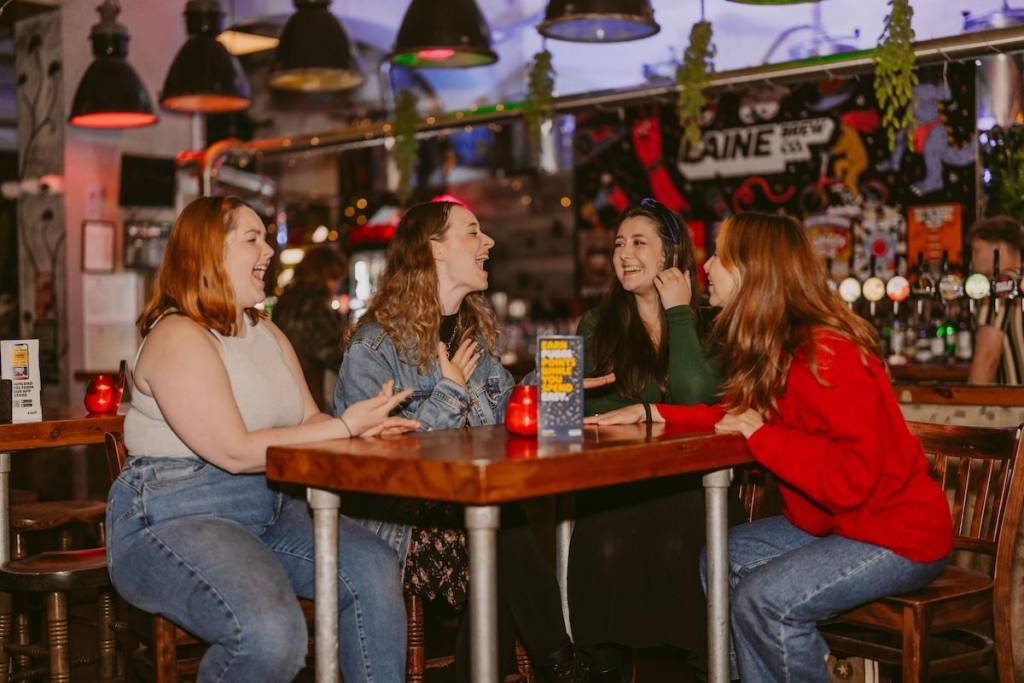4 women sit around a table at a bar talking and laughing