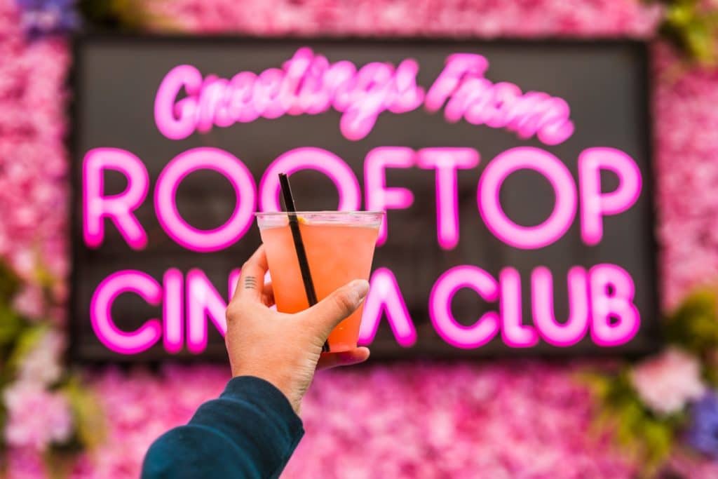 A cocktail pictured in front of a Rooftop Cinema Club neon sign surrounded by flowers