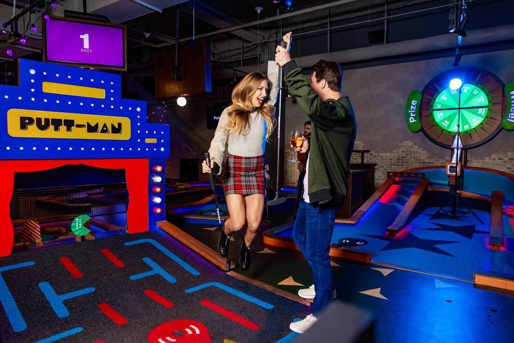 Una pareja disfrutando de una partida de minigolf con toques tecnológicos en Puttshack, sonriendo y sosteniendo los palos en un campo iluminado con luces de neón